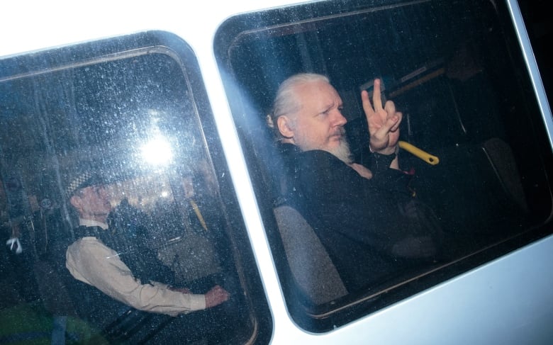 A man with long white hair, tied in a ponytail, with a long white beard, makes a peace sign with his fingers through the window of a van. 