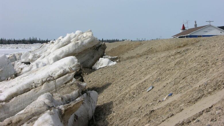 Large chunks of ice creep up the side of an earthen dike with a rooftop in the distance  