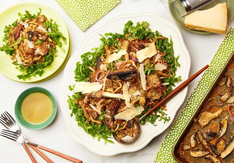 Overhead shot of a serving platter of mushroom and farro salad on a bed of greens. It's sitting on a white table set for dinner.