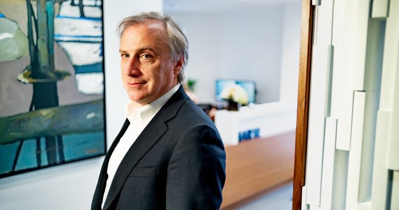 A white man with short grey hair, a dark blazer and a white suit is shown from the shoulders up.