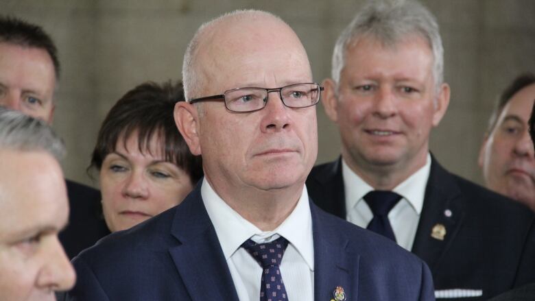 A man with glasses wearing a suit stands in a crowd of people.