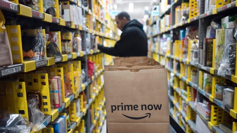 A man wearing a hoodie stands in a warehouse surrounded by tall shelves filled with merchandise. An Amazon Prime bag is in focus in the foreground. 