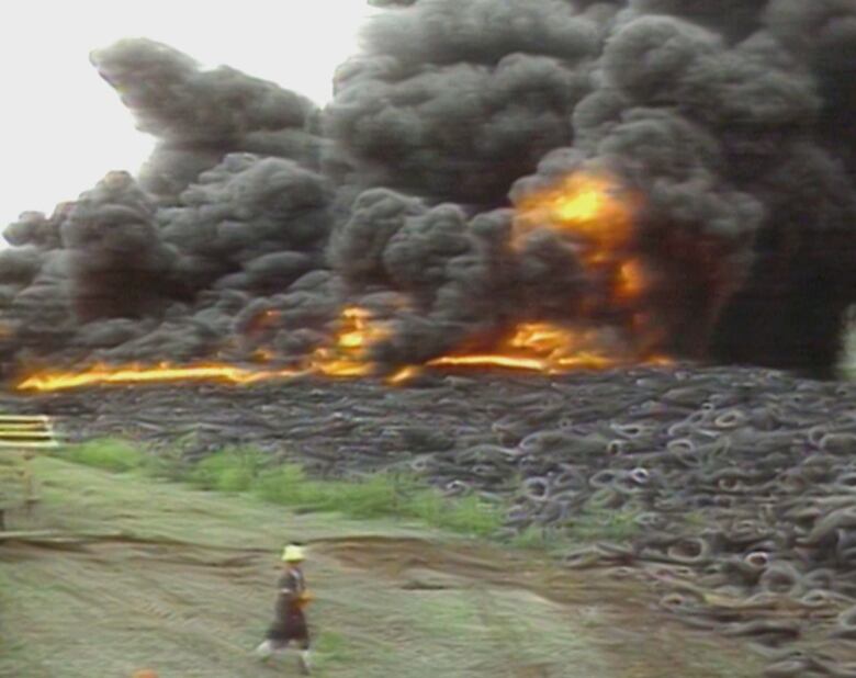 firefighter walks on field in front of fire with thick black smoke emerging from a pile of tires 