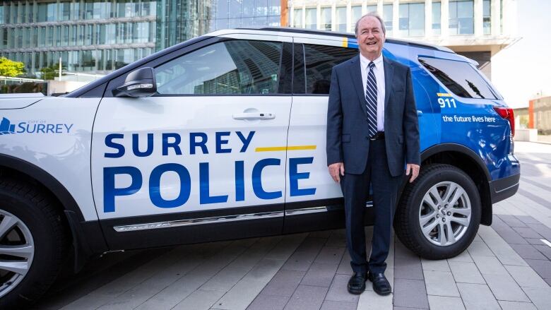 A smiling white man in a dark-blue suit stands beside an SUV emblazoned with the words 