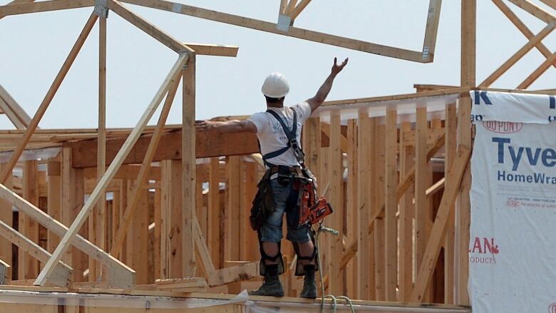 A man standing on the frame of a new home under construction, reaching his hand upwards.