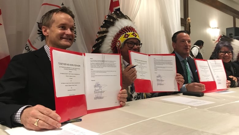 Three men at a table, two wearing suits and one wearing a headdress, hold up red folders with documents inside. 