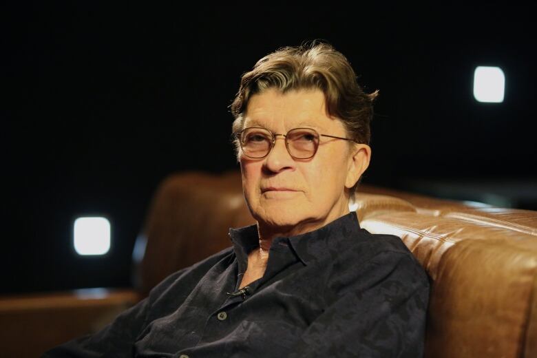 A man wearing glasses sits in a dimly lit theatre.