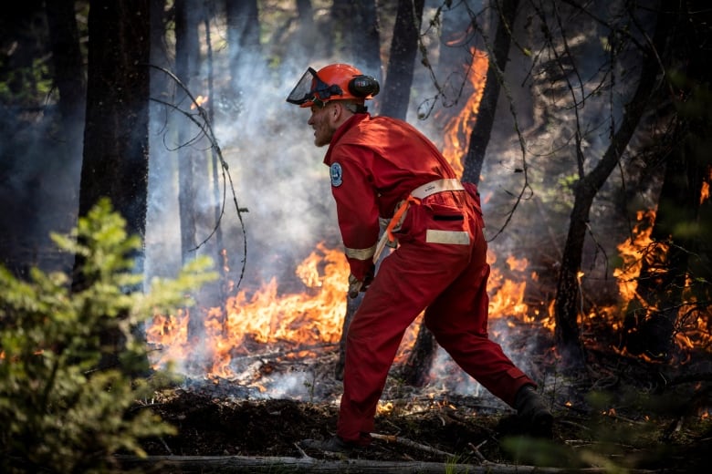 A firefighter in a red jumpsuit with soot on his face stands in smoke near a burning fire in a training exercise.