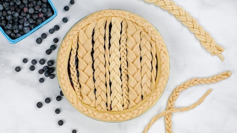 overhead shot of an unbaked blueberry pie on a marble surface. the pie crust is multiple braided pieces of dough. a box of blueberries sits next to the pie dish, with some berries scattered. 