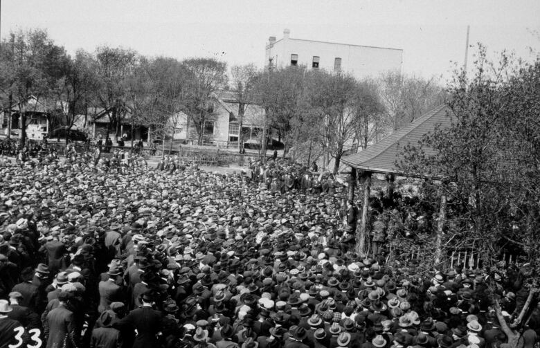 A black-and-white photo taken from above shows a crowd of people, most wearing hats, around a gazebo.