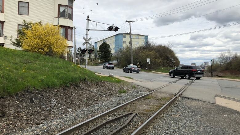 A railroad crossing an urban street in Saint John is shown from the perspective of the tracks. A residential complex is on the left of the photo with beige siding and a green lawn and several cars are driving up the road away from the tracks.