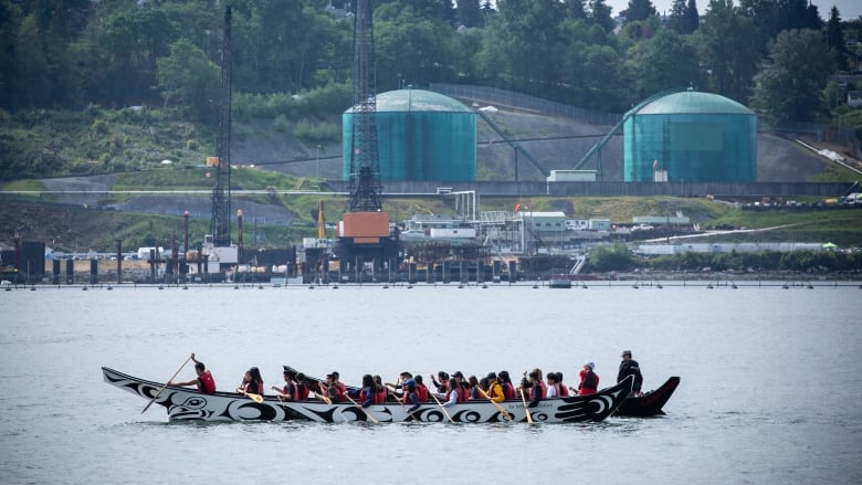 An indigenous canoe is paddled in front of oil storage tanks on the shores of Burrard Inlet.