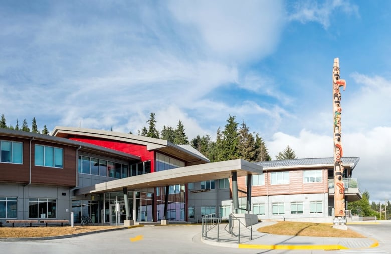 The front entrance of Haida Gwaii Hospital is a modern building with a large rectangular awning and a totem pole out in front of it.