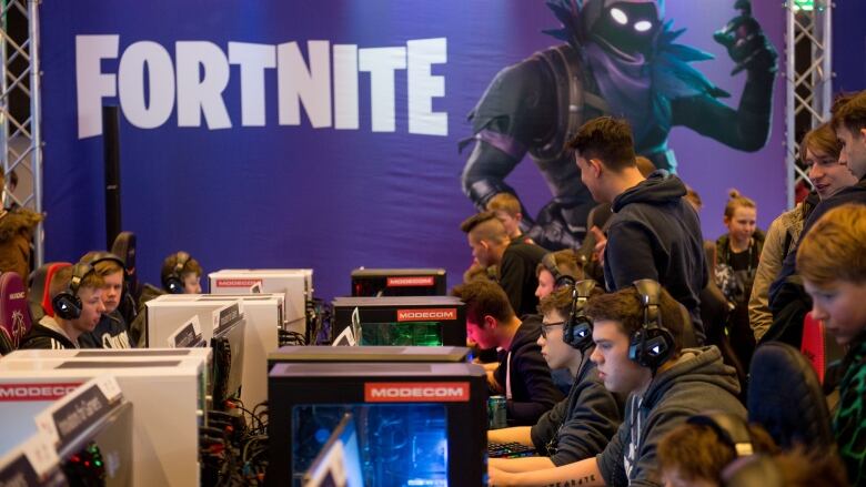 A bunch of young people sit in front of computers, with the poster of Fortnite in the background.