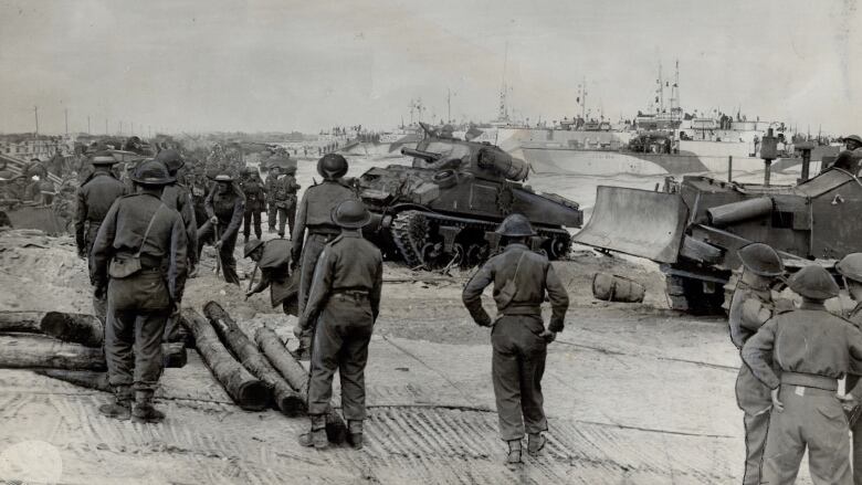 Second World War soldiers walk along a beach during the Normandy Invasion as ships land and tanks and bulldozers are unloaded.