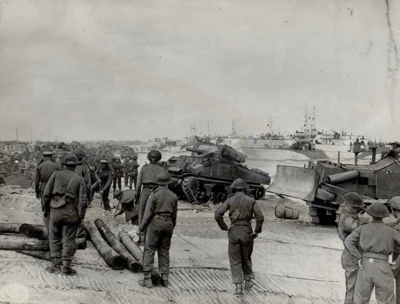 Second World War soldiers walk along a beach during the Normandy Invasion as ships land and tanks and bulldozers are unloaded.