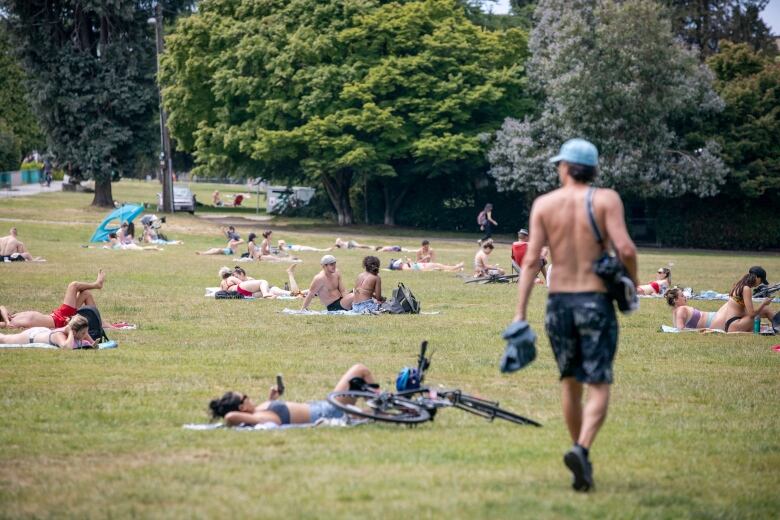 A range of people lay down on towels in a grassy area. Some of them are shirtless.