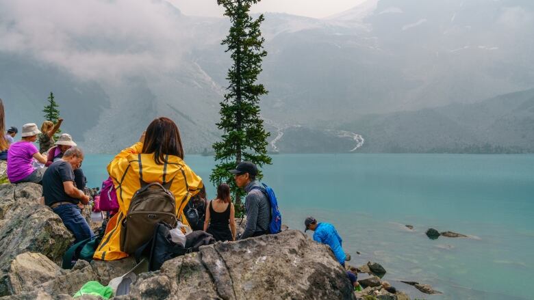 A person sits on a rock looking out at a turquoise lake. They have shoulder-length brown hair and wear a yellow jacket and brown backpack. Other people scattered along the rocks, look out at the lake.