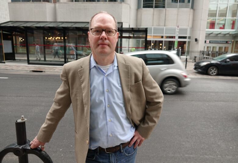 A man in a blazer and collared shirt stands on a downtown street as a car passes behind him.