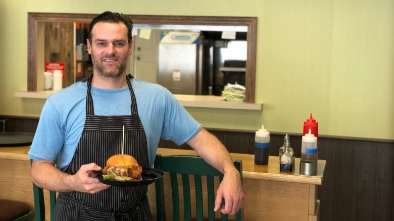 A man in an apron holds a plate containing a massive fried chicken sandwich.