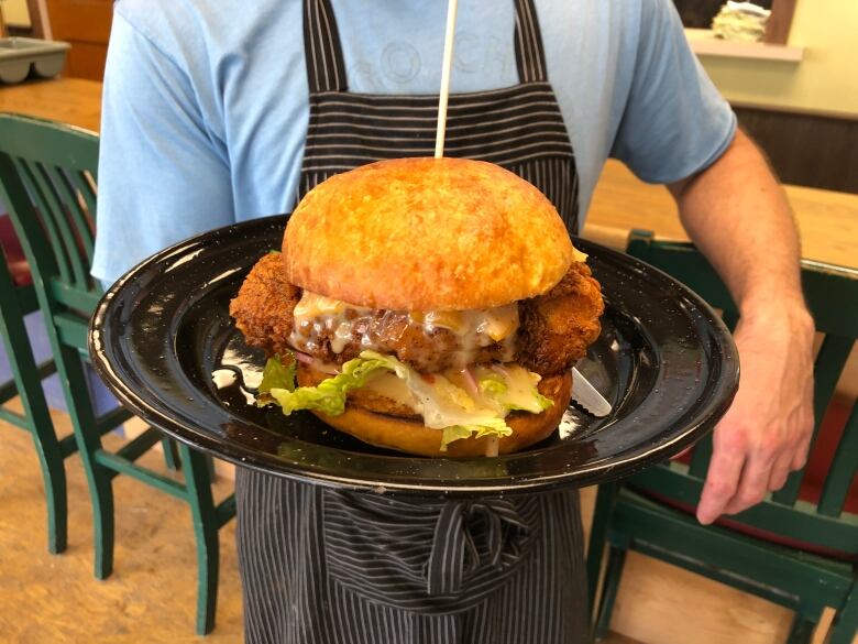 A person in an apron holding up a plate with a massive fried chicken sandwich.