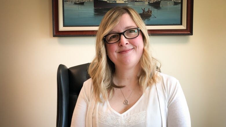 A woman with glasses, blonde hair and a white blouse and sweater sits at a desk and smiles.