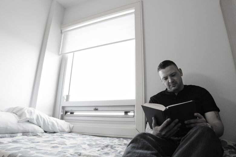 A man reads a book while sitting on a bed next to an open window in a black and white photo.