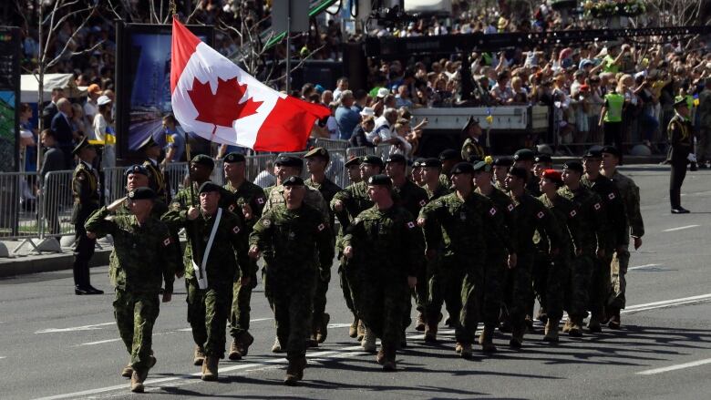 A group of soldiers march down a street on a sunny day with crowds and a Canadian flag in the background.