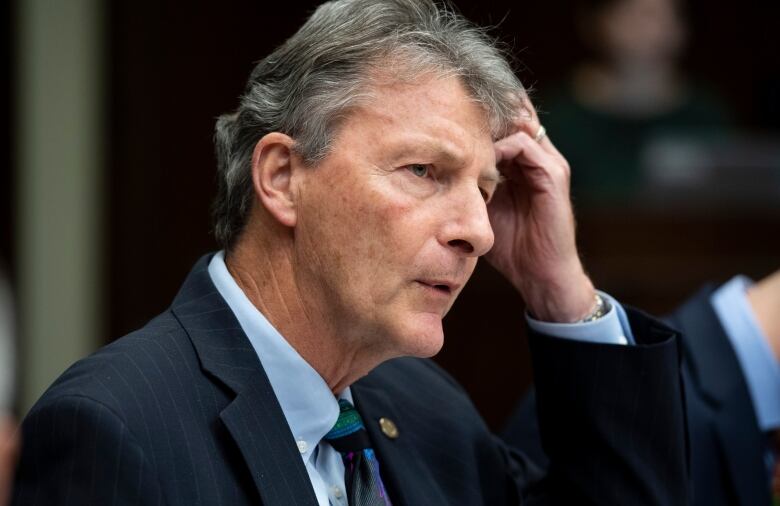 A man wearing a suit and tie scratches his head.