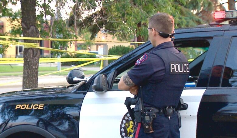 A Calgary police officer stands beside a police car near yellow crime scene tape.