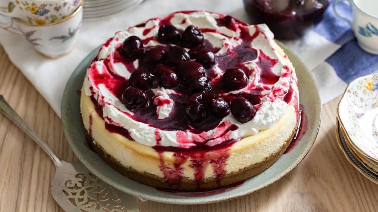 Overhead shot of a cheesecake sitting on a serving plate on a wooden table. The cheesecake is topped with cherry topping. 