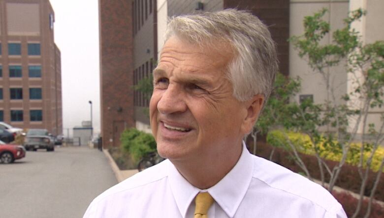 A man with grey hair in a pink shirt speakers to reporters outside on a city street.