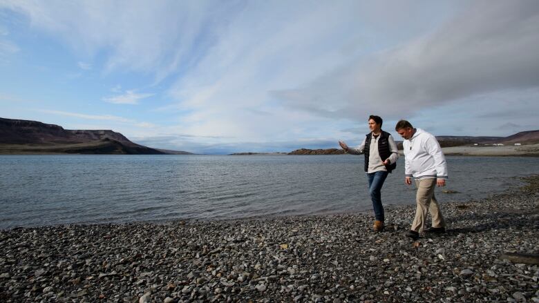 Two men walk on a gravel beach in the High Arctic with tundra covered mountains in the background. 