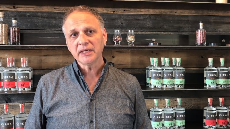 A man looks slightly off-camera, standing in front of a shelf holding bottles of clear alcohol with red and turquoise labels and the logo for the distillery Cirka.