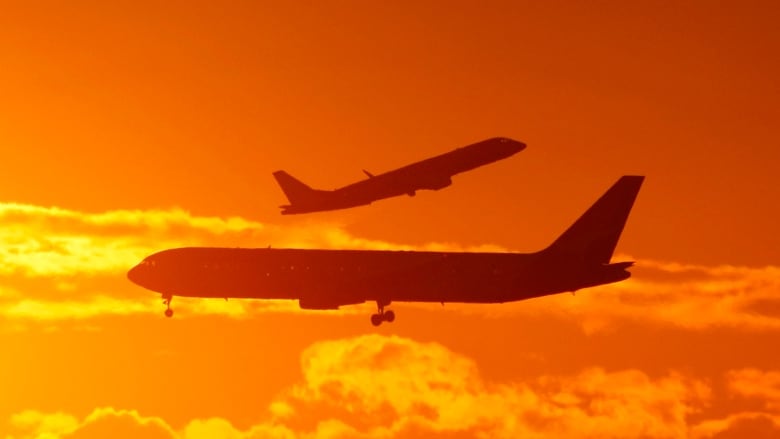 The silhouettes of two planes can be seen passing each other in an orange sky. 