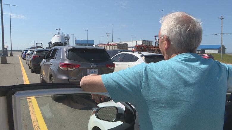 A man stands at an open car door with several other vehicles ahead of him. A large passenger vessel is in the background with its nose cone open to allow vehicles on. 