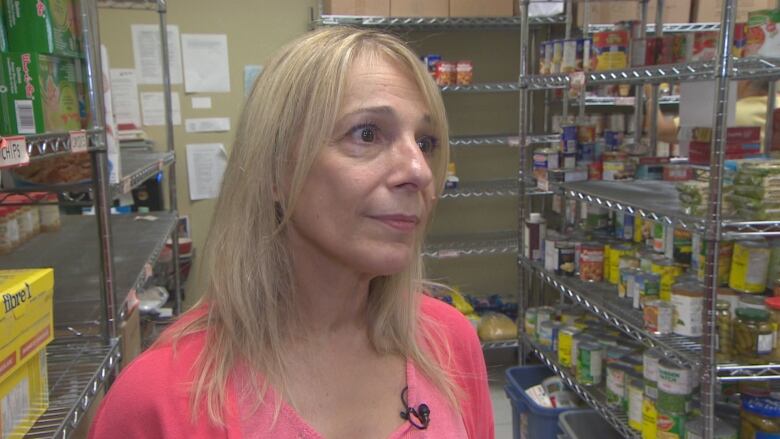 A middle-aged blonde woman is shown in a pink shirt from the shoulders up. She is standing by stocked food shelves.