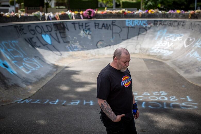 Carson's father Aron Crimeni stands inside a skateboard bowl at the park where his son overdosed. He is standing amidst memorial messages written in chalk, including a 1-800 number for a suicide helpline. He wears black jeans and a black shirt and has tattoos on his bare arms and a slightly greying beard and shortcut hair.