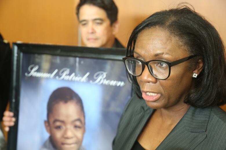 A woman speaking. There's a photo of a young child behind her that reads 