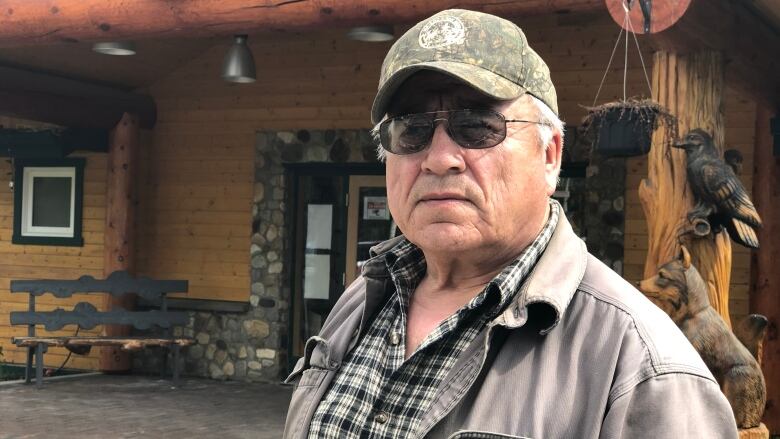 Man wearing a hat and sunglasses standing infront of a log cabin.