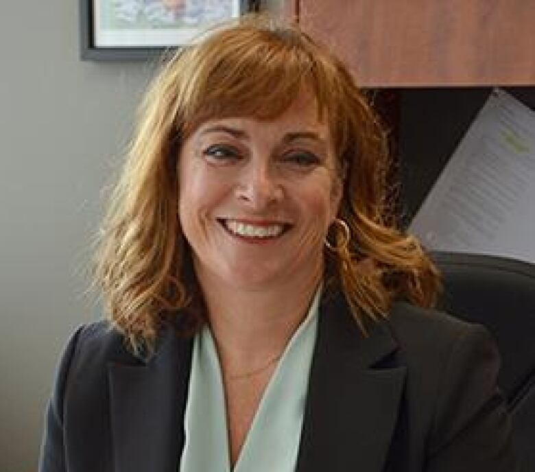 A head shot of a woman sitting behind a desk in an office. She has medium-length wavy red hair with bangs and is wearing a dark suit over a lighter v-neck blouse. 