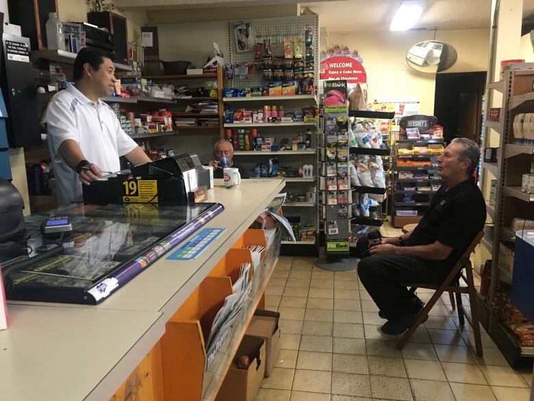 An older Chinese man stands behind a counter in a small shop, speaking with another man sitting on a chair on the shop floor.