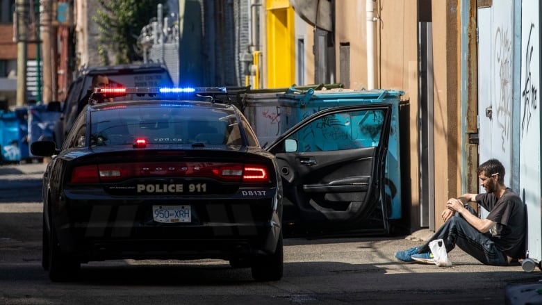 A police car with one door open is visible next to a man sitting in an alley.