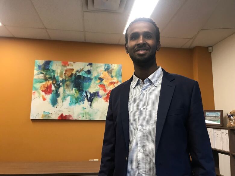 A man in a collared shirt and blazer stands in a room with a painting behind him