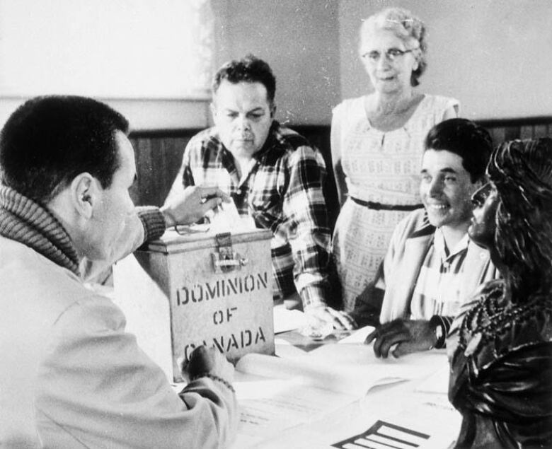 Black-and-white photo shows a man placing a ballot into a Dominion of Canada voting box while other individuals look on. 