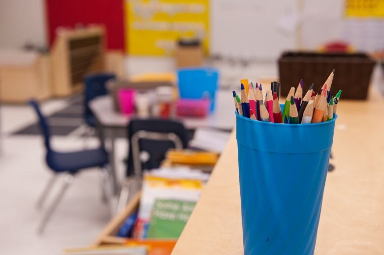 Coloured-pencils are in a bright blue cup. It sits on a table in a classroom. 