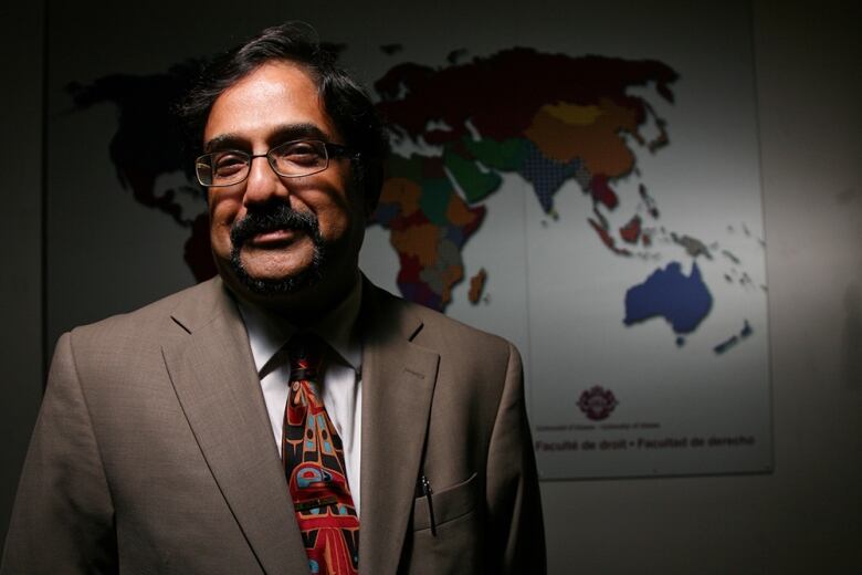 A man in a brown suit stands in front of a map of the world.