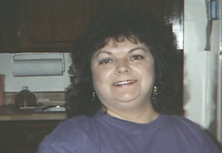 A woman with shoulder-length dark curly hair smiles at the camera, she's wearing a dark blue or purple sweatshirt.