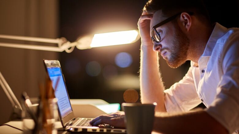 A man wearing glasses sits at a desk at night and stares at a laptop screen, stressed.