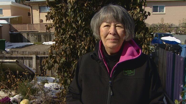 A woman with gray hair smiles in front of a garden. She appears to be in a back yard. She is wearing a black coat. 